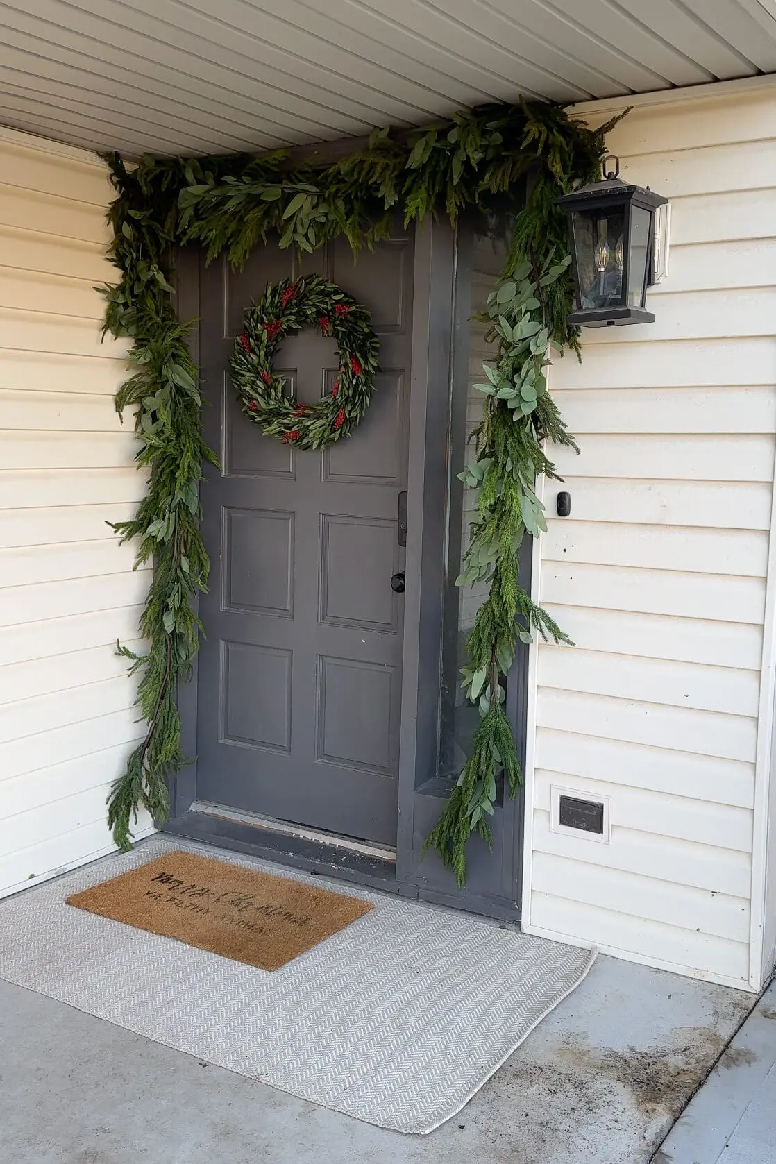 A front porch decorated for the holidays with lush greenery garland framing a gray door and a festive wreath featuring red accents. The entrance is complemented by a neutral outdoor rug and a natural coir doormat with a cheerful Christmas greeting.